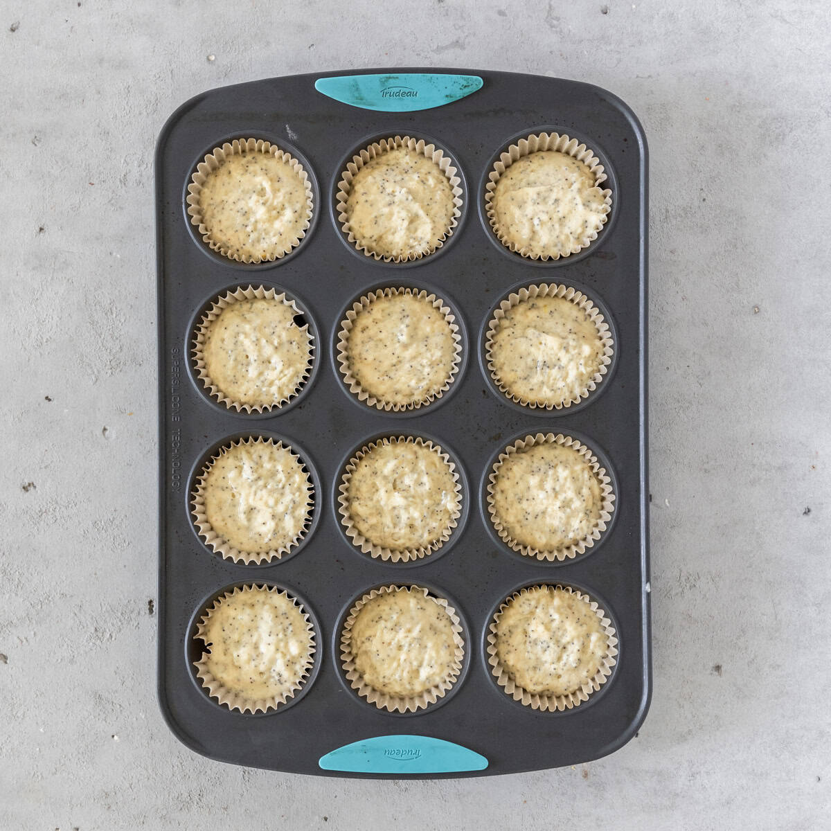 the lemon poppy seed muffins in a muffin tin before being baked on a grey background.