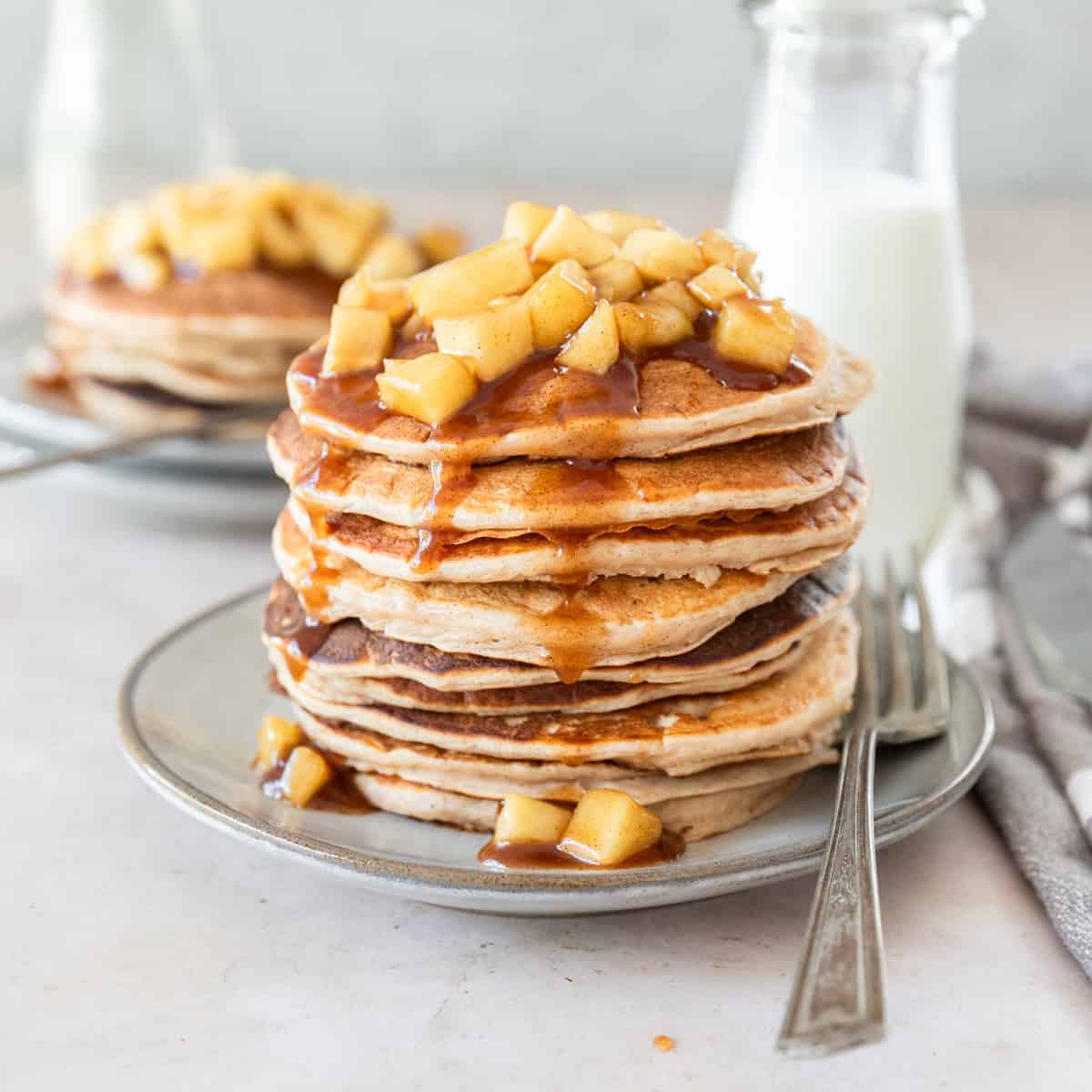 a large stack of fluffy apple cinnamon pancakes on a grey plate with a metal fork beside it.