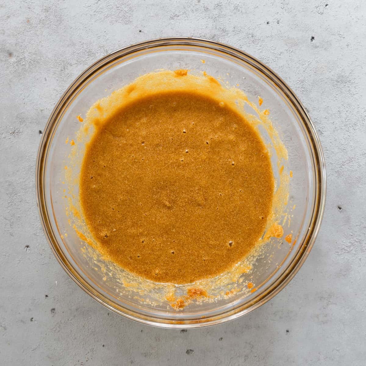 all of the wet ingredients and sugar combined in a glass bowl on a grey background.
