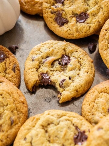 a tray of soft and chewy pumpkin chocolate chip cookies on a silver tray. One cookie has a bite out of it.