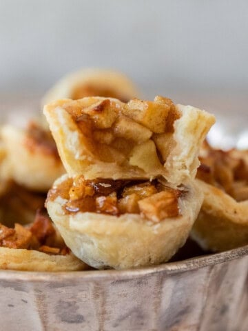 several mini apple pies in a silver dish. Two are stacked on top of each other and one has a bite out of it.