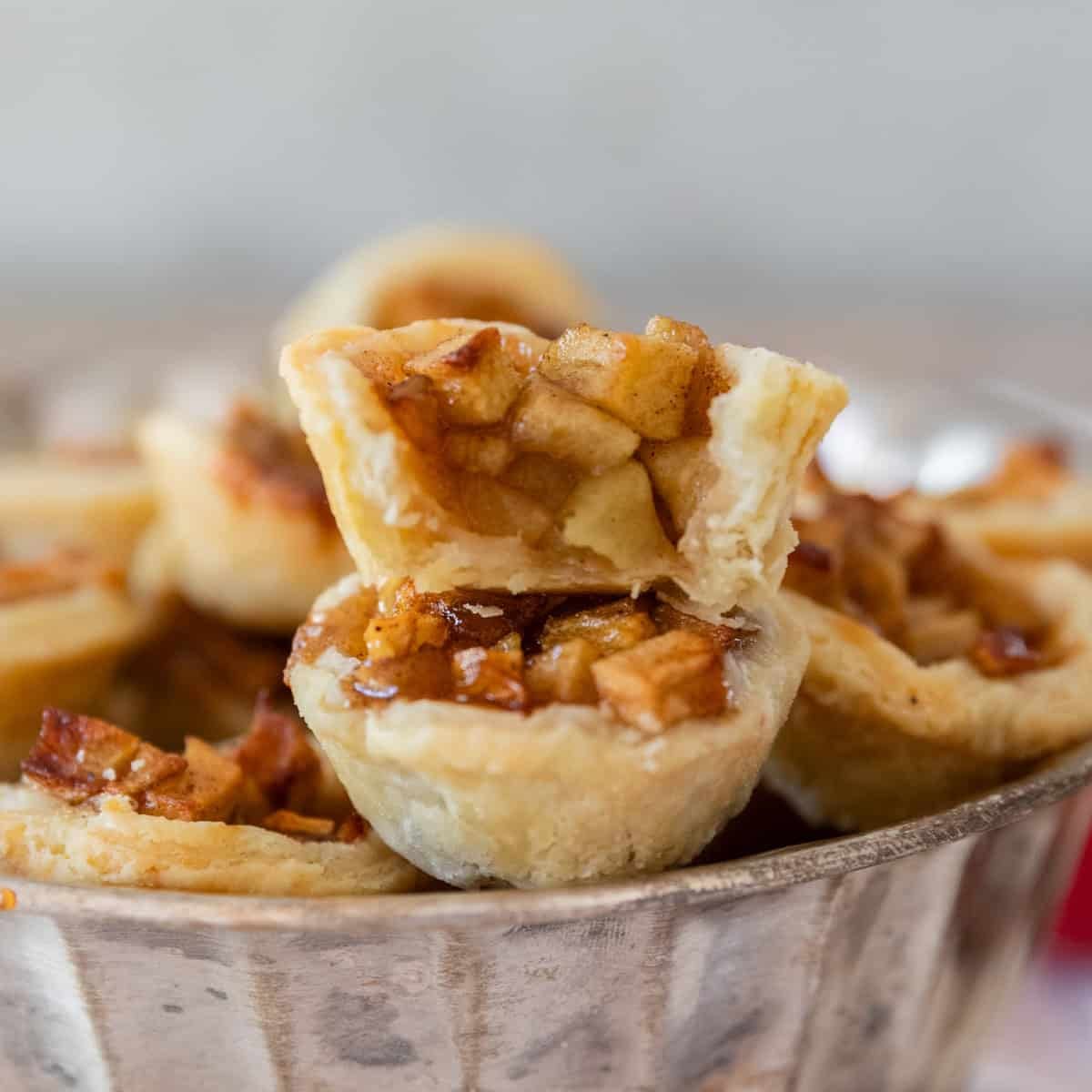 several mini apple pies in a silver dish. Two are stacked on top of each other and one has a bite out of it.