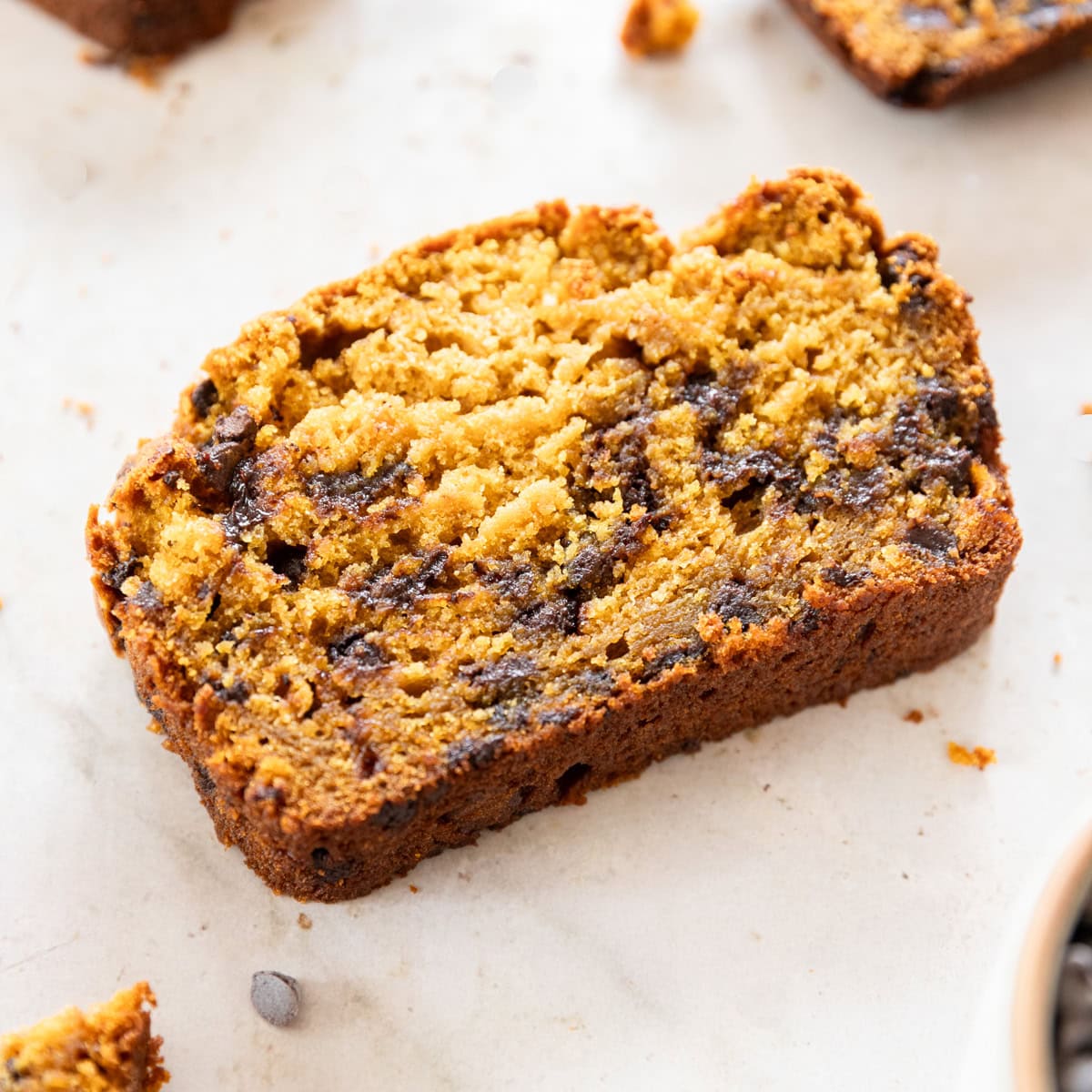 a slice of pumpkin chocolate chip bread with other slices around it on a tan background.