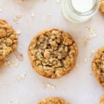 several soft and chewy oatmeal cookies on a tan background next to a glass of milk with oats scattered around.